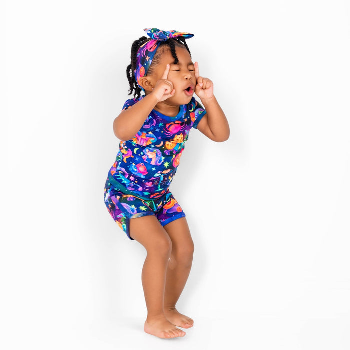 A young child models the Jammie Session Cosmic Cuddles Zodiac Short Pajama Set with a matching headband, standing barefoot and playfully posing against a plain white background.