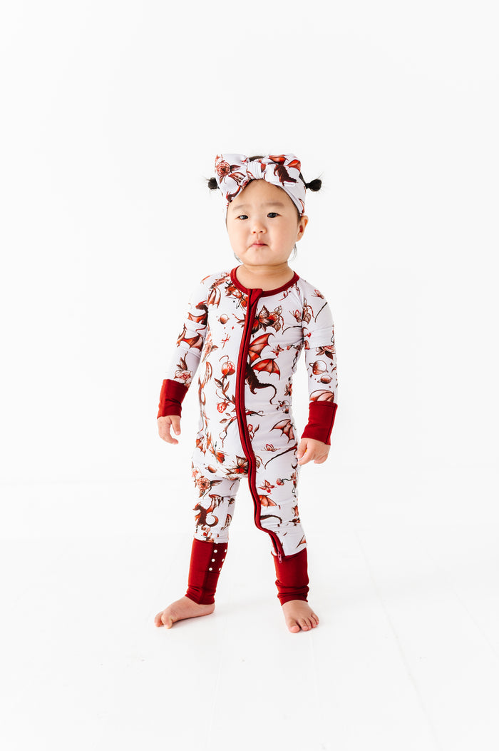 A toddler wears the Jammie Session Dreaming with Dragons Romper, a white and red bamboo pajama set with dragon print and matching headwrap, standing barefoot against a plain white background.