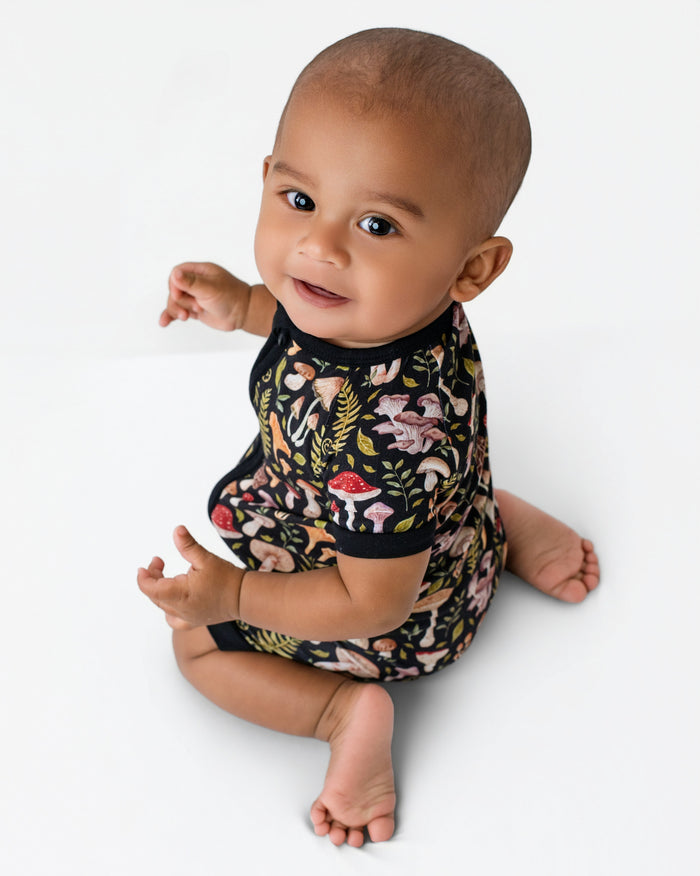A baby sits on a white background, wearing the Jammie Session Mystic Mushrooms Short Sleeve Romper, which features a colorful botanical and mushroom print. The baby looks up at the camera with a slight smile.