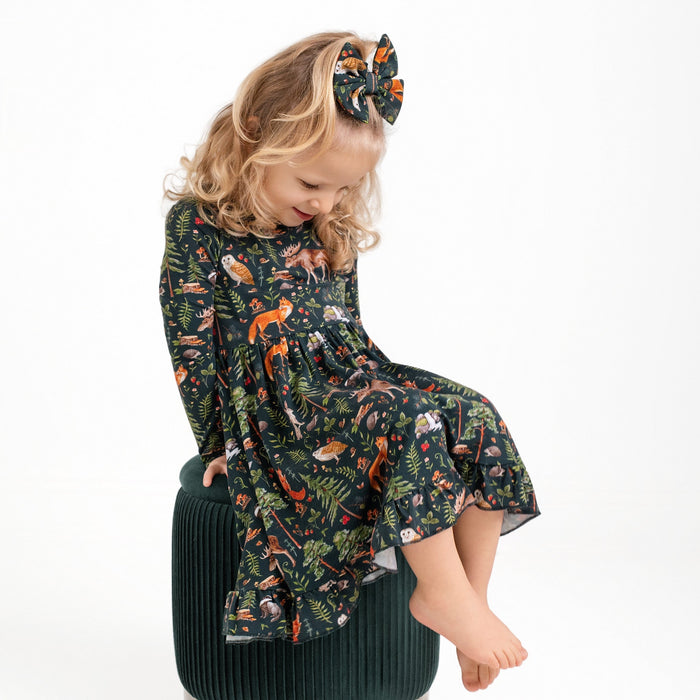 A young blonde girl sits barefoot on a green stool, wearing the Jammie Session Wood You Be My Friend Long Sleeve Big Kid Twirl Dress with colorful animal and plant patterns, plus a matching hair bow.