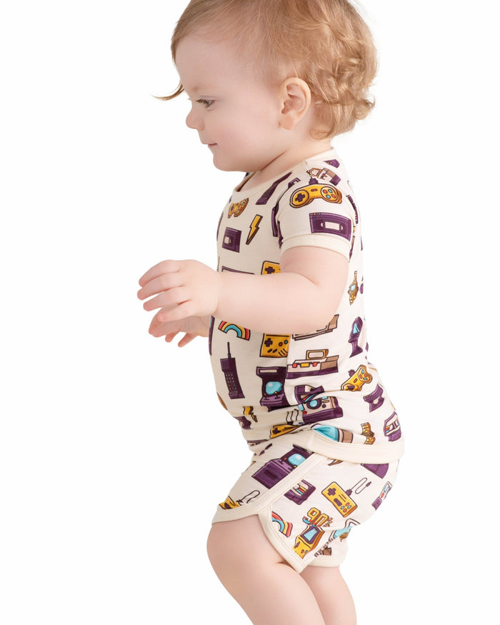 A toddler stands against a plain white background wearing the Jammie Session 90's Retro Short Pajama Set‚Äîbreathable, hypoallergenic bamboo baby clothes featuring colorful retro prints with matching short sleeves and shorts.