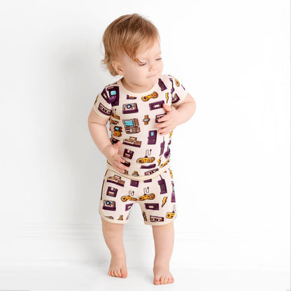 A toddler stands barefoot against a white wall, wearing the Jammie Session 90's Retro Short Pajama Set featuring a breathable matching shirt and shorts with colorful vintage electronics and gadget prints.