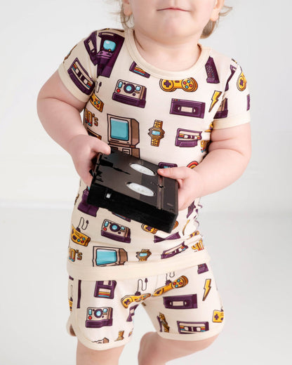 A toddler in the Jammie Session 90's Retro Short Pajama Set‚Äîbreathable bamboo pajamas with fun TV, watch, game console, and cassette tape prints‚Äîholds a VHS tape. Perfect for sensitive skin.