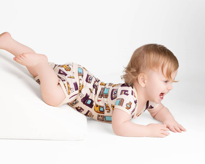 A baby in a Jammie Session 90's Retro Short Pajama Set crawls up a white wedge cushion against a plain white background.