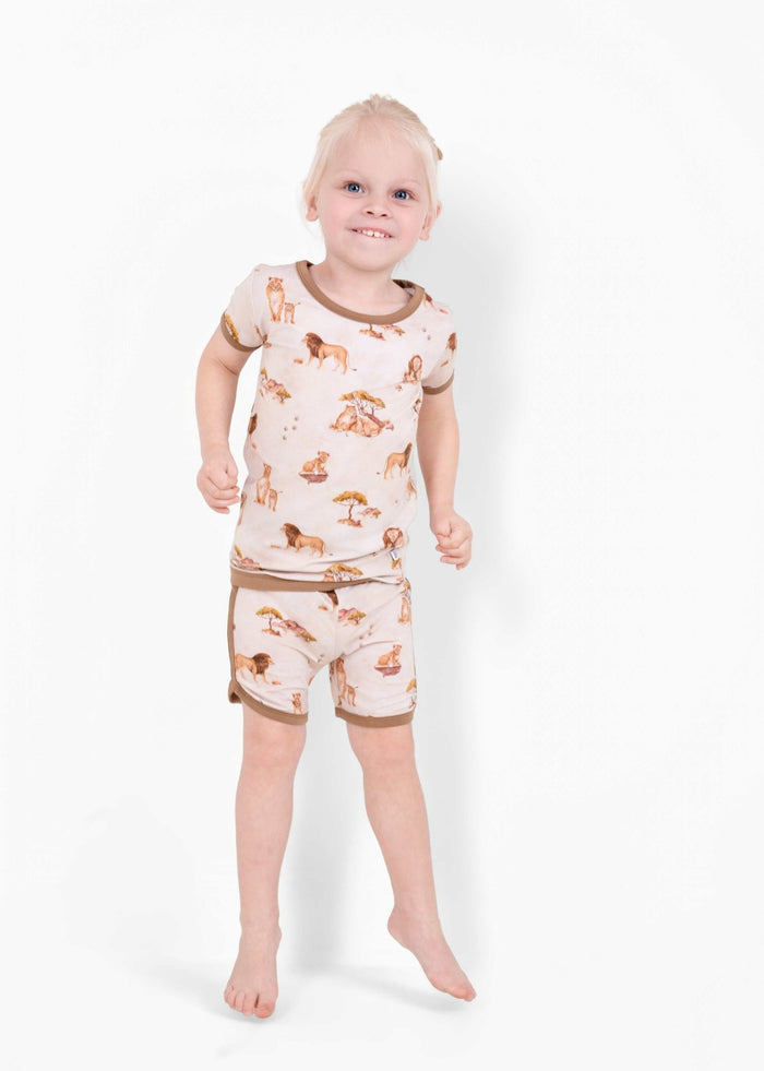 A young child with light hair wears the Jammie Session Ari (Lion) TENCEL‚Ñ¢ Modal Short Pajama Set, featuring buttery soft, matching animal print top and shorts, standing barefoot against a plain white background.