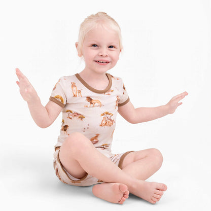 A young blond child sits cross-legged, smiling with hands up, wearing Jammie Session's Ari (Lion) TENCEL‚Ñ¢ Modal Short Pajama Set‚Äîhypoallergenic, buttery soft pajamas featuring animal prints and brown trim.