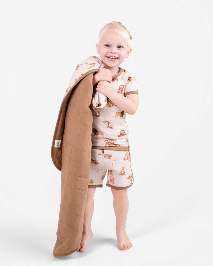 A smiling young child in the Ari (Lion) TENCEL‚Ñ¢ Modal Short Pajama Set by Jammie Session holds a tan quilted blanket made from buttery soft, hypoallergenic fabric against a plain white background.
