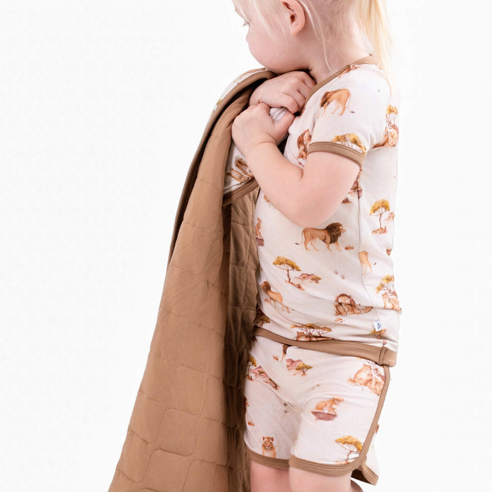 A young child wears the Jammie Session Ari (Lion) TENCEL‚Ñ¢ Modal Short Pajama Set‚Äîfeaturing buttery soft, animal-print fabric‚Äîwhile holding a brown quilt against a plain white background.