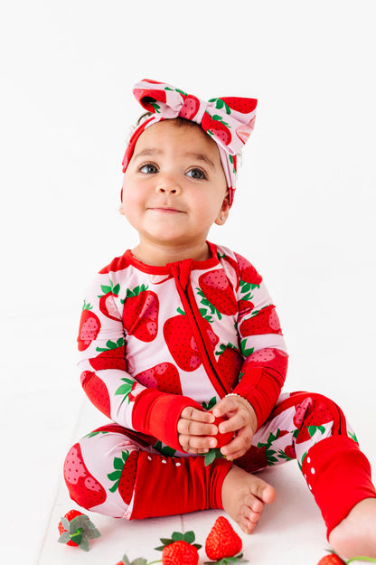 A baby wears the Jammie Session Berry Sweet (Strawberry) Romper and matching headband, sitting on the floor holding a strawberry, with more strawberries scattered around them on a white background.