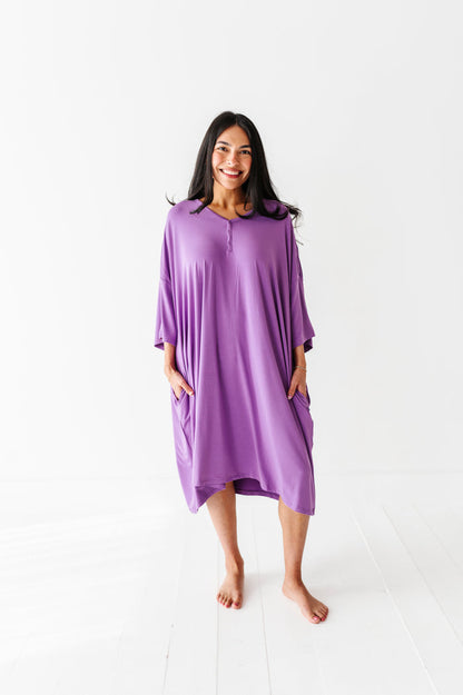 A woman stands barefoot on a white floor, wearing the Jammie Session Block Party Solid Women's Muumuu Lounge Tee (ONE SIZE FITS XS - XXL) in purple with pockets, set against a plain white background.