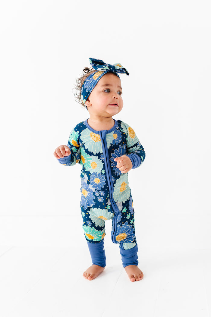 A toddler wears the Jammie Session Bluepsie Daisy Romper‚Äî a blue floral bamboo long-sleeve romper with convertible footies and matching headband‚Äî while standing against a plain white background.
