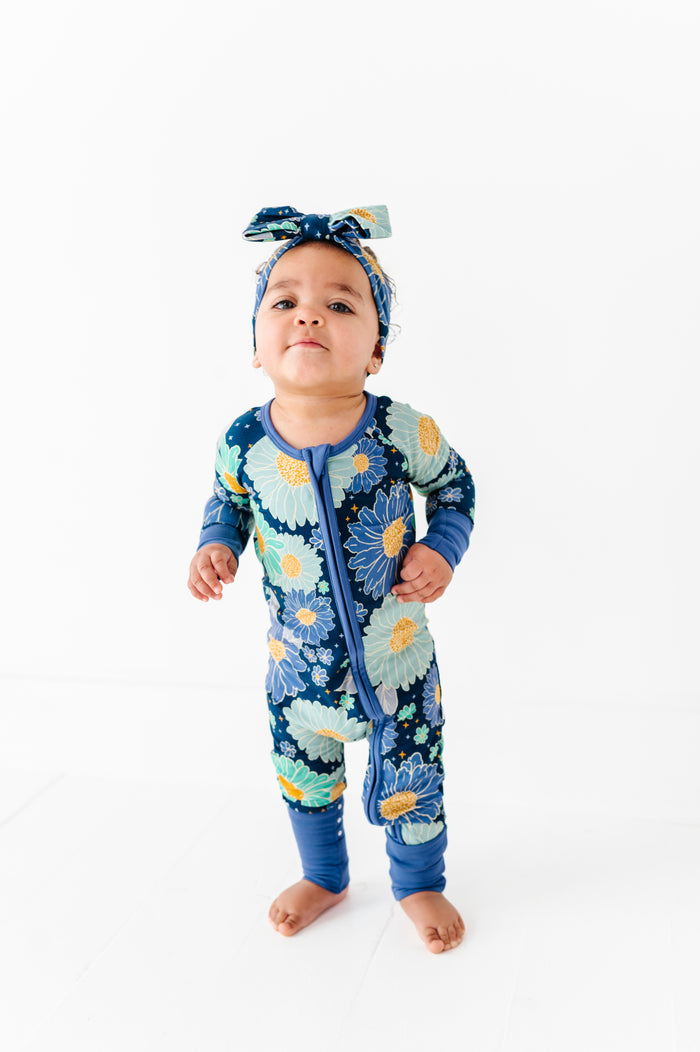 A baby wears the Jammie Session Bluepsie Daisy Romper with long sleeves, convertible footies, a matching headband, and a double zipper for easy changes, standing barefoot on a white floor against a plain white background.