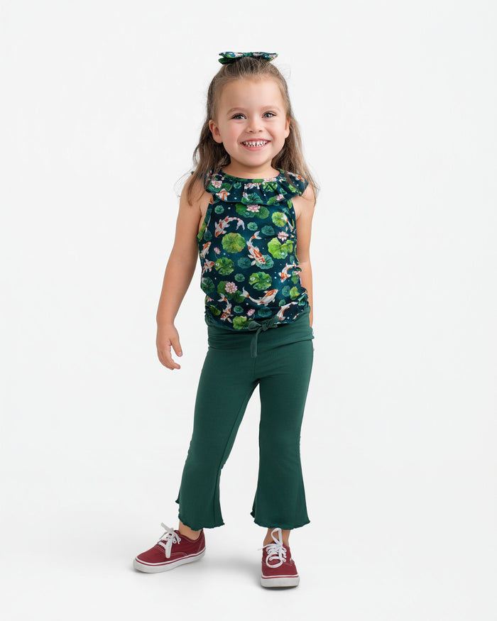 A young girl smiles in the Jammie Session Bundle of Koi Bloomy Crop Set‚Äîfeaturing a sleeveless floral top and green flared pants‚Äîpaired with red sneakers and a coordinating hair bow against a plain white background.