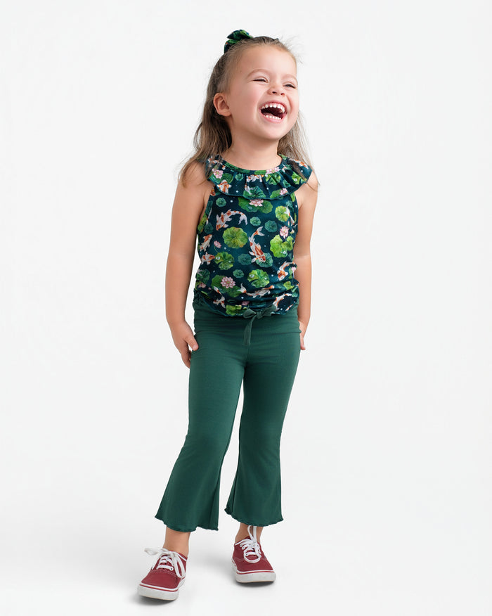 A young child smiles in the Jammie Session Bundle of Koi Bloomy Crop Set, featuring a green floral sleeveless top and matching pants, paired with red sneakers and a green hair tie, against a plain white background.