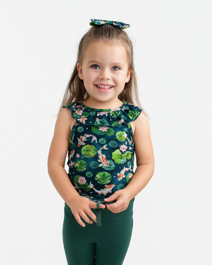 A young child with long brown hair wears the Jammie Session Bundle of Koi Bloomy Crop Set‚Äîfeaturing a matching floral top, hair bow, and dark green pants‚Äîstanding and smiling against a plain white background.