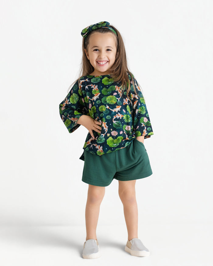 A young girl stands smiling in the Jammie Session Bundle of Koi Comfy Cloud Short Set, featuring a matching green floral top, bow headband, green shorts, and light slip-on shoes against a white background.