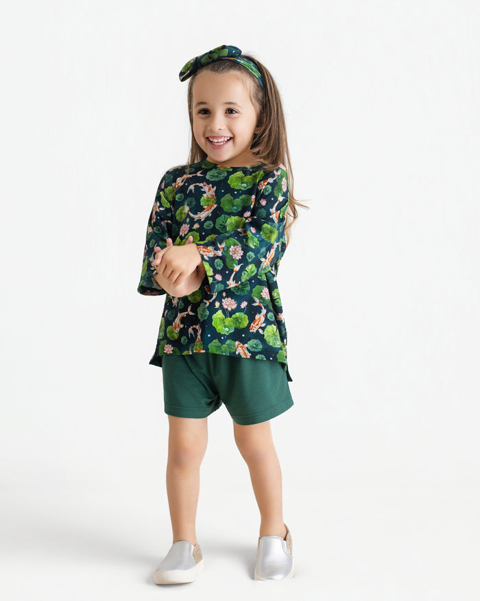 A young girl smiles in the Jammie Session Bundle of Koi Comfy Cloud Short Set‚Äîfeaturing a green floral long-sleeve top, matching shorts, and a floral headband‚Äîwith silver slip-on shoes, set against a plain white background.