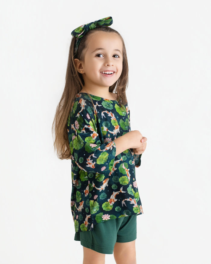 A young girl smiles in the Jammie Session Bundle of Koi Comfy Cloud Short Set‚Äîdark green floral top with matching headband and green shorts‚Äîstanding against a plain white background.