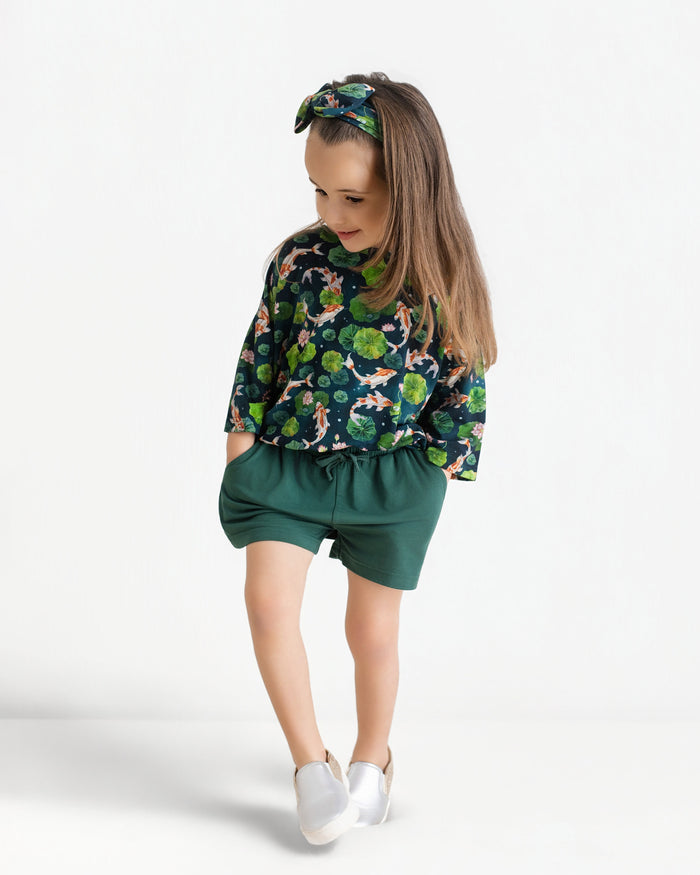 A young girl wears the Jammie Session Bundle of Koi Comfy Cloud Short Set‚Äîgreen-patterned top, matching shorts, and headband‚Äîwith white shoes, standing with hands in pockets against a plain white background.