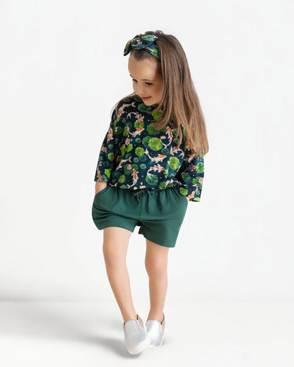 A young girl wears the Jammie Session Bundle of Koi Comfy Cloud Short Set‚Äîgreen-patterned top, matching shorts, and headband‚Äîwith white shoes, standing with hands in pockets against a plain white background.