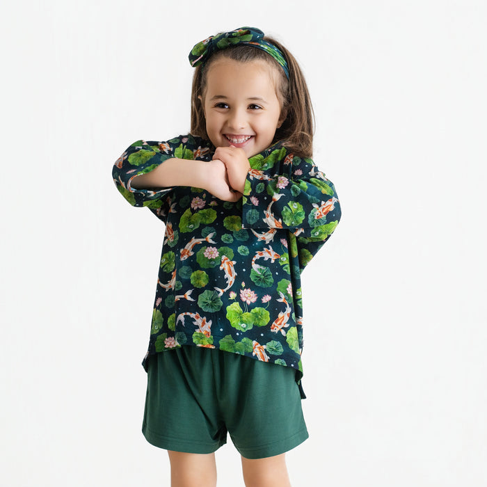 Young child smiles with arms crossed, wearing the Jammie Session Bundle of Koi Comfy Cloud Short Set‚Äîfeaturing a koi fish and lily pad pattern shirt, dark green shorts, and matching headband‚Äîagainst a plain white background.