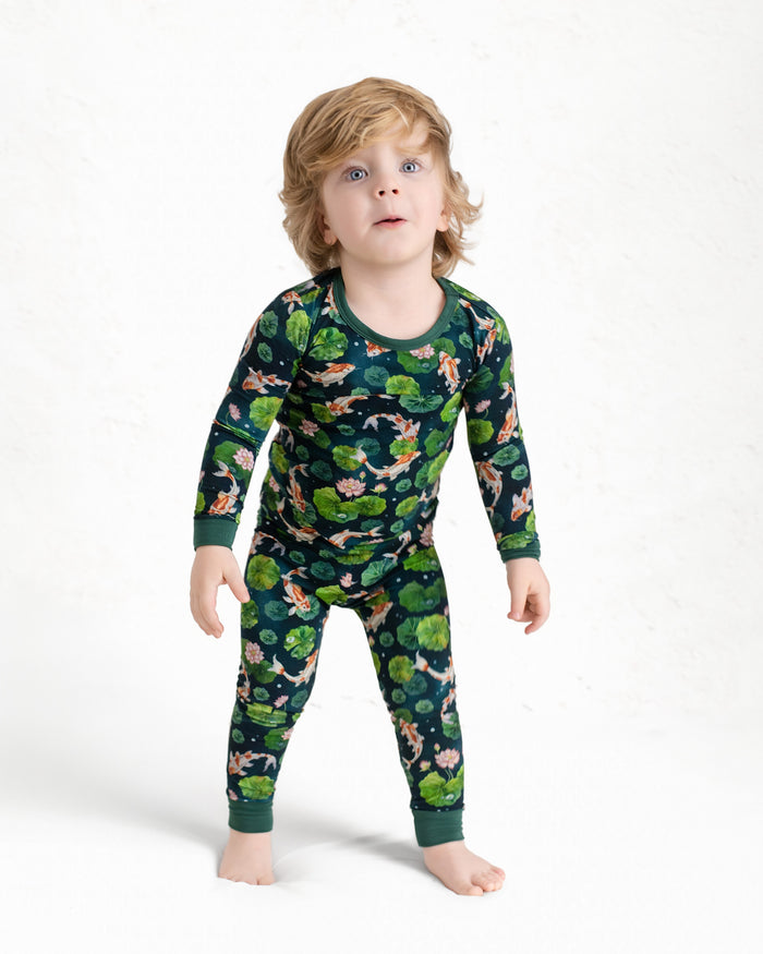 A young child with light brown hair stands barefoot against a white background wearing Jammie Session‚Äôs Bundle of Koi Long Sleeve PJ's‚Äîhypoallergenic kids pajamas in dark green with a nature and animal print.