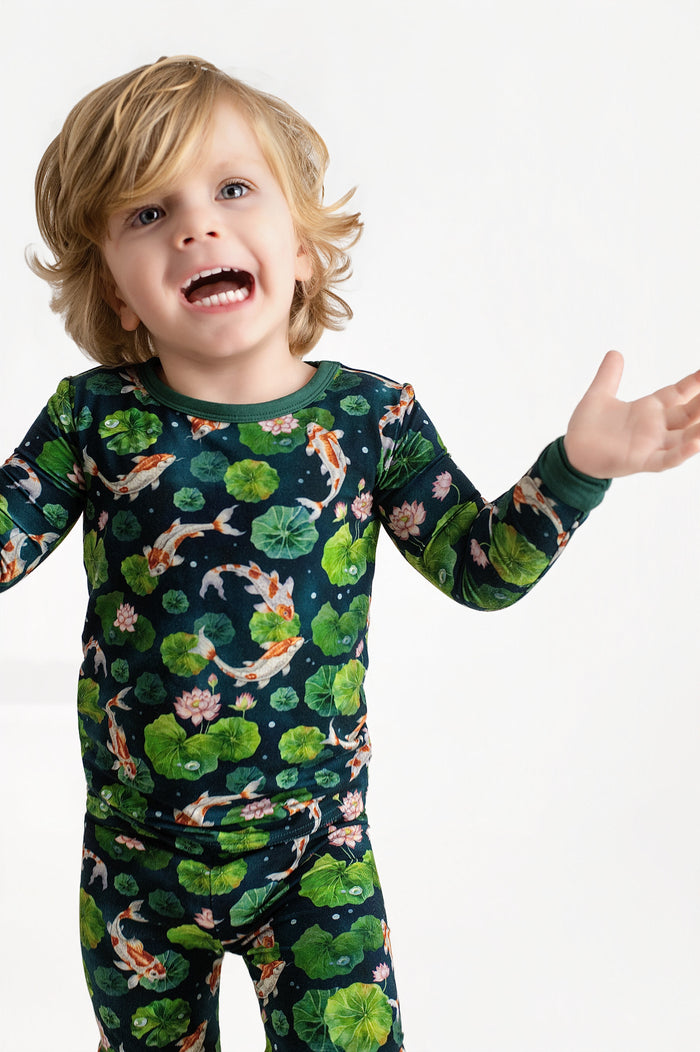 A young child with blonde hair wears Jammie Session‚Äôs Bundle of Koi Long Sleeve PJ's, featuring hypoallergenic fabric and a koi fish and lily pad print, standing against a plain white background with arms raised and mouth open.