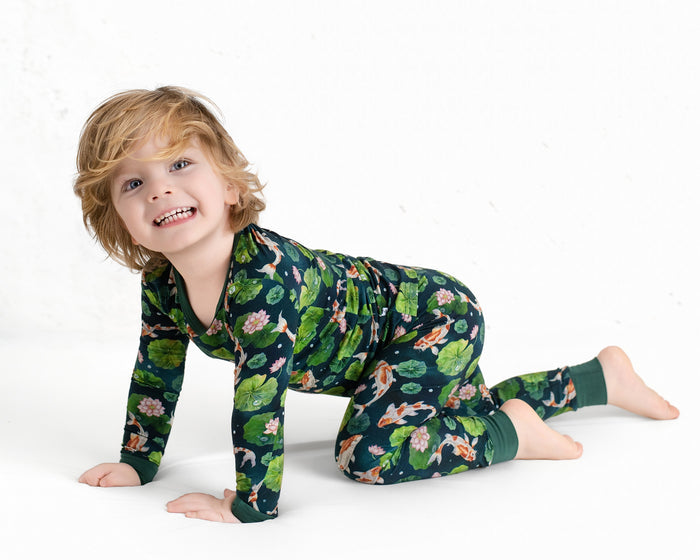 A young blond child, smiling at the camera, crawls on a white background while wearing Jammie Session‚Äôs Bundle of Koi Long Sleeve PJ's featuring a dark green color with floral and animal patterns.