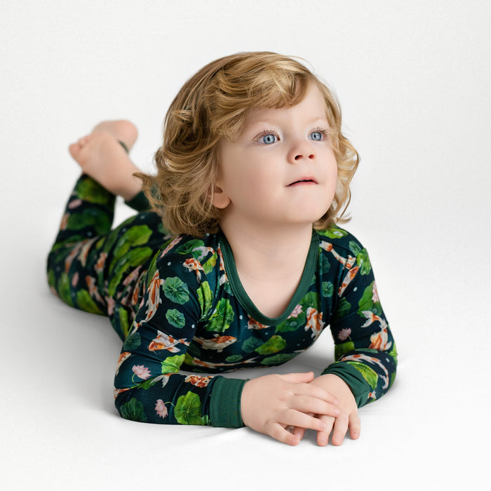 A young child with curly blond hair and blue eyes lies on their stomach in Jammie Session's Bundle of Koi Long Sleeve PJ‚Äôs, featuring a floral and animal print, against a plain white background.