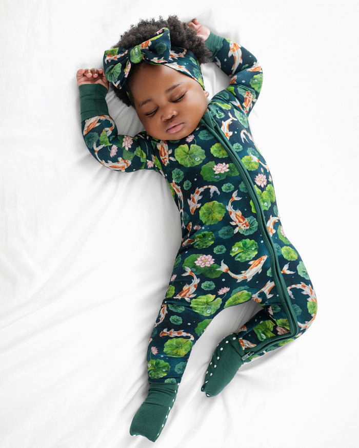 A baby wears the Jammie Session Bundle of Koi Romper‚Äîa dark green floral and koi fish patterned convertible footed romper with a matching headband‚Äîwhile sleeping on a white surface.
