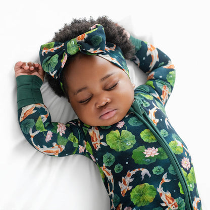 A baby in the Jammie Session Bundle of Koi Romper‚Äîa long sleeve bamboo romper with a koi fish and lily pad pattern‚Äîwears a matching headband and sleeps on a white surface with arms raised.
