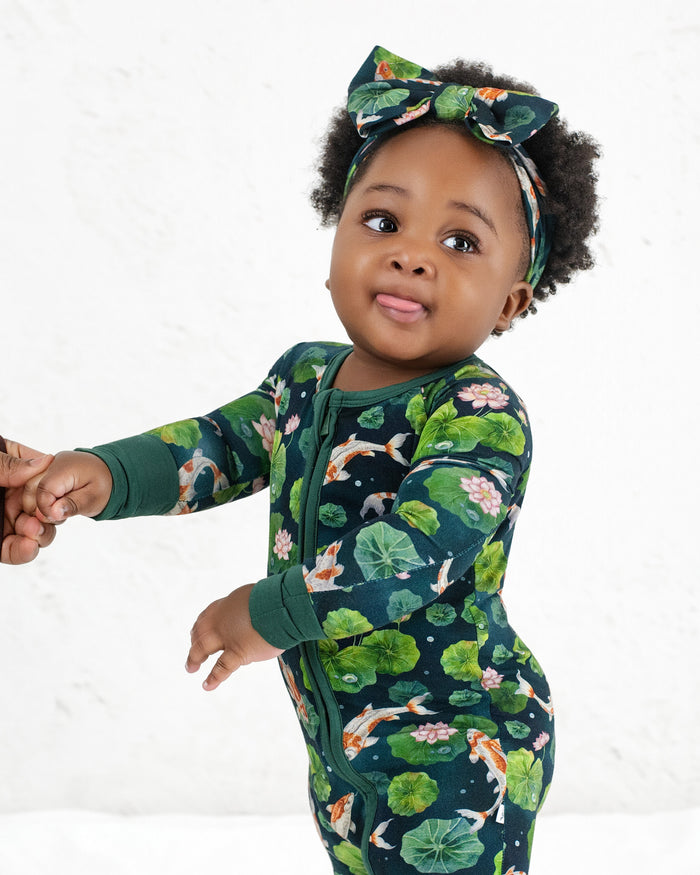 A toddler wears the Jammie Session Bundle of Koi Romper with a matching headband, holds an adult‚Äôs hand, and looks ahead with their tongue slightly out.