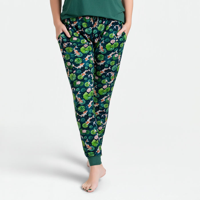 A woman wears Jammie Session's Bundle of Koi Women's Jogger Pants‚Äîdark green with a vibrant floral and animal print‚Äîhands in pockets, standing barefoot against a white background.