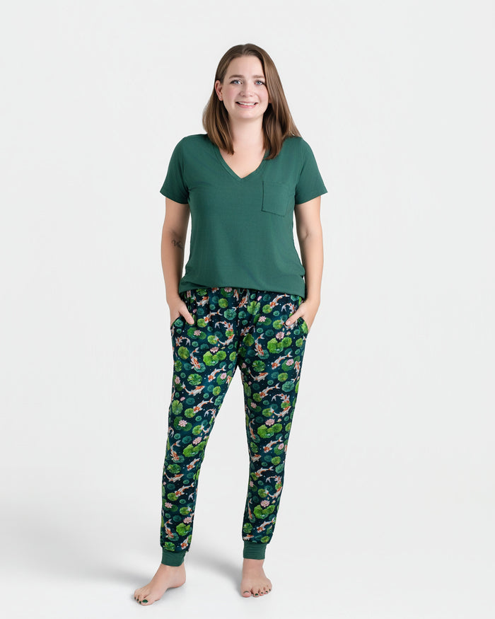 A woman stands barefoot in a Jammie Session Bundle of Koi Women's Solid Short Sleeve T-shirt and patterned pajama pants, hands in pockets. The moisture-wicking fabric keeps her comfortable all day against a plain white background.