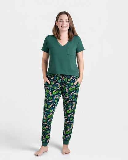 A woman stands barefoot in a Jammie Session Bundle of Koi Women's Solid Short Sleeve T-shirt and patterned pajama pants, hands in pockets. The moisture-wicking fabric keeps her comfortable all day against a plain white background.