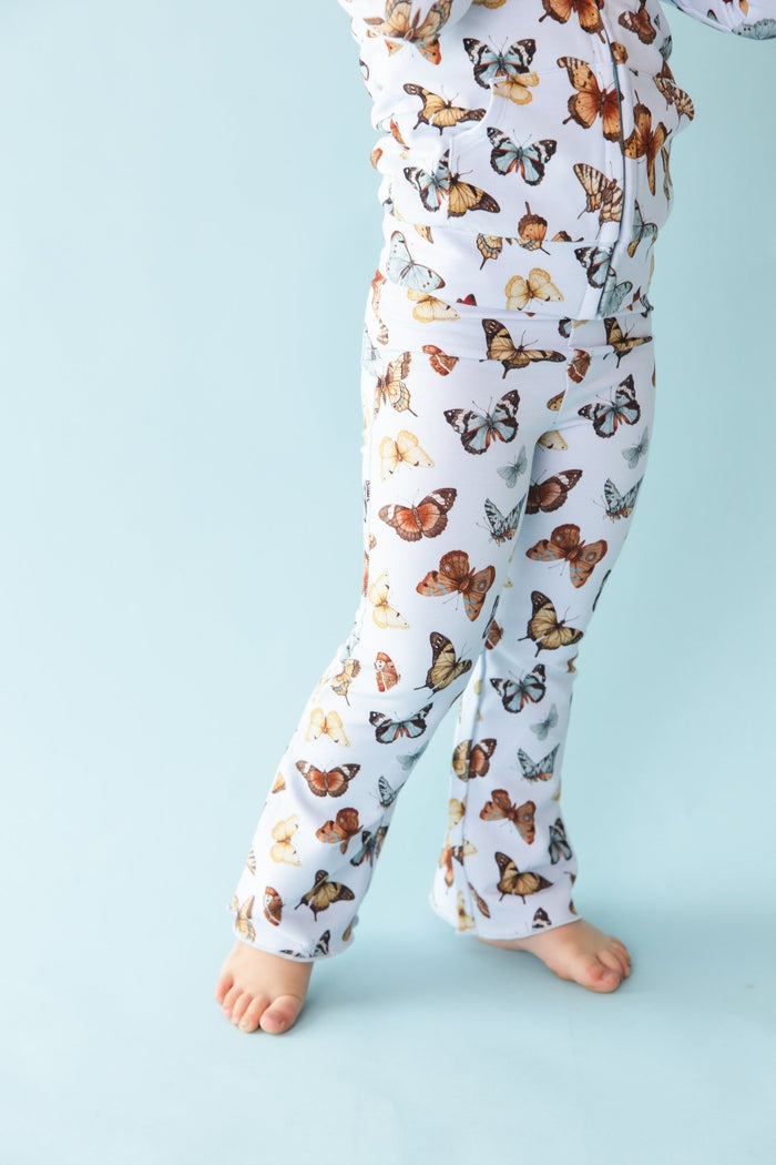A child is wearing Jammie Session‚Äôs Butterfly Kisses Kids' Flare Pants‚Äîbreathable white pajamas with a colorful butterfly print, shown barefoot against a light blue background, lower body only visible.