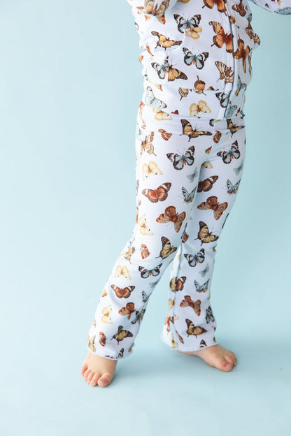 A child is wearing Jammie Session‚Äôs Butterfly Kisses Kids' Flare Pants‚Äîbreathable white pajamas with a colorful butterfly print, shown barefoot against a light blue background, lower body only visible.