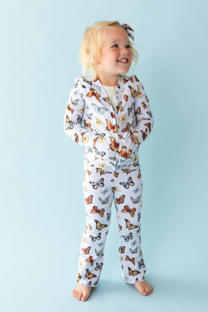 A young child smiles barefoot against a light blue background, wearing Jammie Session‚Äôs Butterfly Kisses Kids' Flare Pants‚Äîhypoallergenic and crafted from a soft bamboo cotton blend.