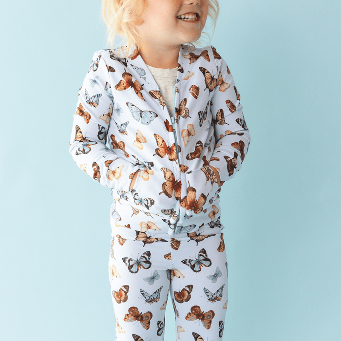 A young child smiles with hands in pockets against a light blue background, wearing the Jammie Session Butterfly Kisses Kids' Jacket (runs small, please size up) and matching pants set with a butterfly print.