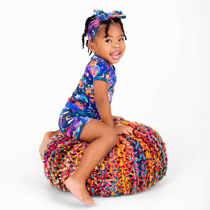 A young child in the Jammie Session Cosmic Cuddles Zodiac Short Pajama Set and a matching headband smiles while sitting on a multicolored knitted pouf against a white background.