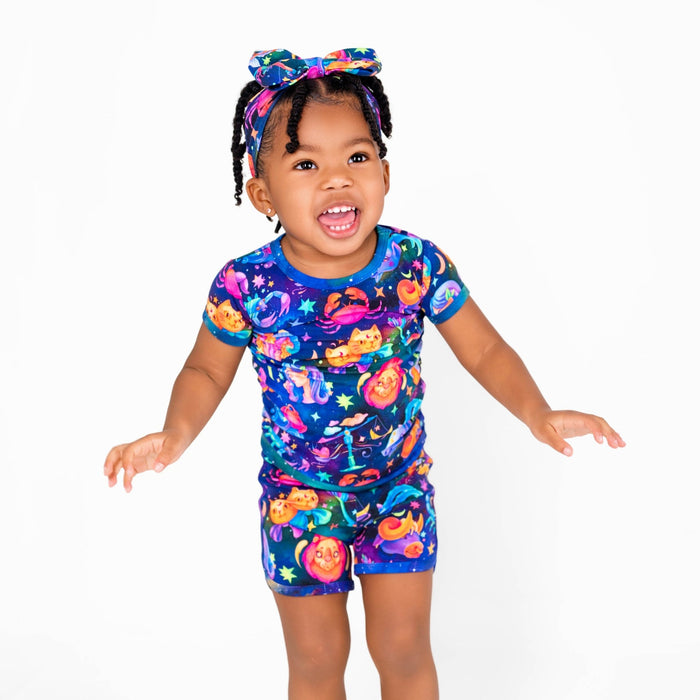 A young child wears the buttery soft, hypoallergenic Cosmic Cuddles Zodiac Short Pajama Set by Jammie Session in blue, paired with a colorful Halloween-themed headband, standing against a plain white background.