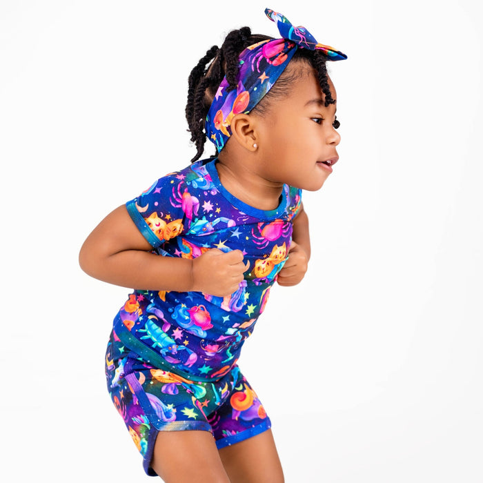 A young girl poses against a white background, wearing Jammie Session's Cosmic Cuddles Zodiac Short Pajama Set‚Äî buttery soft, hypoallergenic bamboo baby clothes with a colorful, space-themed design and matching headband.