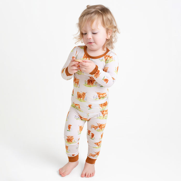 A young child in Jammie Session‚Äôs Cozy Cattle Long Sleeve PJ's‚Äîwhite, hypoallergenic bamboo pajamas with a brown and orange farm animal print‚Äîstands barefoot holding an object against a plain white background.