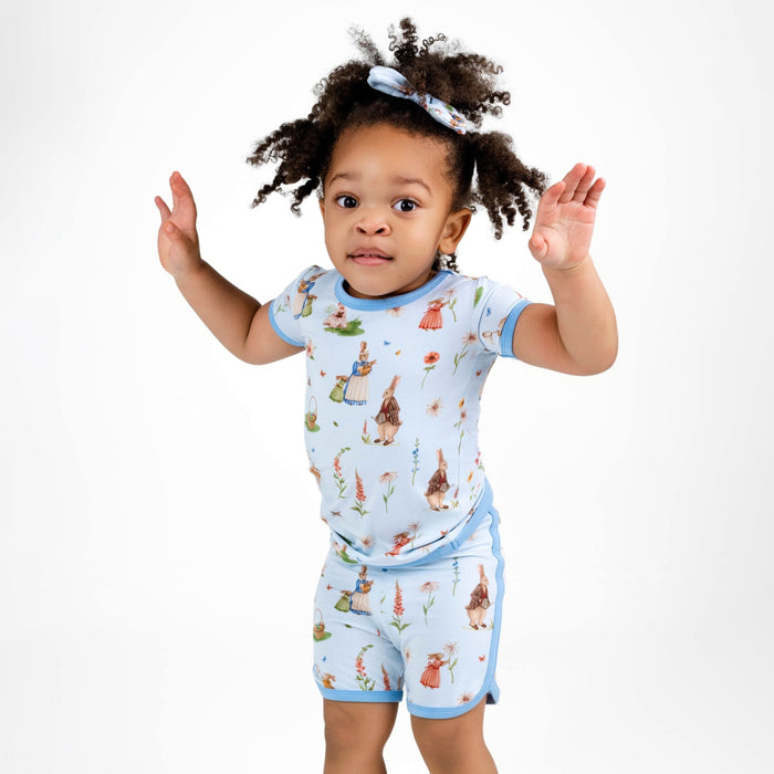A young child in the Jammie Session ‚ÄúDon‚Äôt Worry, Be Hoppy (Rabbit)‚Äù blue bamboo short pajama set with playful illustrations stands with raised arms against a plain white background.