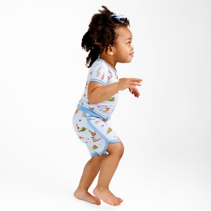 A young child with curly hair stands barefoot against a plain white background, wearing the Jammie Session ‚ÄúDon‚Äôt Worry, Be Hoppy (Rabbit)‚Äù Short Pajama Set featuring light blue and colorful prints.
