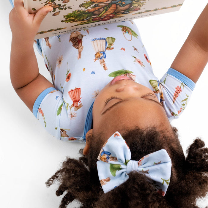 A child with curly hair and a matching bow reads while lying down, cozy in the Jammie Session ‚ÄúDon‚Äôt Worry, Be Hoppy (Rabbit)‚Äù short pajama set‚Äîsoft bamboo fabric that‚Äôs gentle on sensitive skin, hypoallergenic, and moisture-wicking.