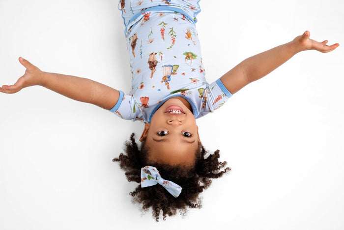 A young child smiles and lies on their back with arms outstretched, wearing the Jammie Session ‚ÄúDon‚Äôt Worry, Be Hoppy (Rabbit)‚Äù short pajama set and matching headband, photographed from above on a white background.
