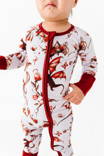 A toddler models the Jammie Session Dreaming with Dragons Romper‚Äîa white and red bamboo outfit featuring dragon and floral patterns‚Äîagainst a white background.