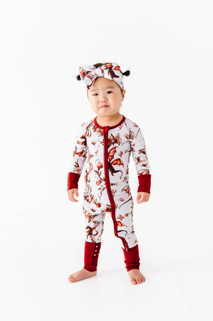 A toddler faces forward against a white background, wearing the Jammie Session "Dreaming with Dragons Romper," a red and white printed long sleeve baby romper, paired with a matching headband.