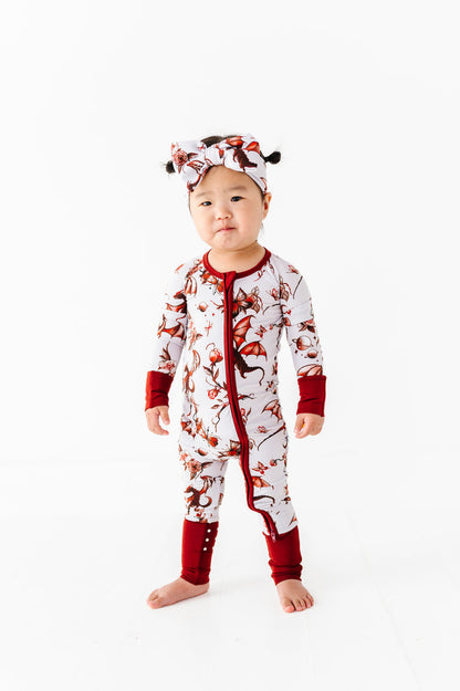 A toddler faces forward against a white background, wearing the Jammie Session "Dreaming with Dragons Romper," a red and white printed long sleeve baby romper, paired with a matching headband.